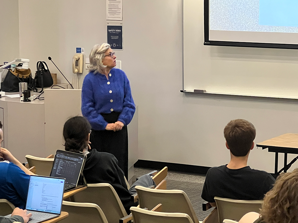 Professor Pamela Troyer, wearing a blue sweater, stands at the front of a classroom giving a lecture. Several students are seated facing her, some with laptops open. The classroom has a projector screen and a lectern with various items on it.