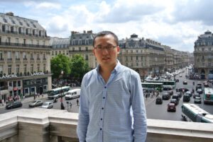 Dr. Andong Ma standing on a balcony with a european city center behind him