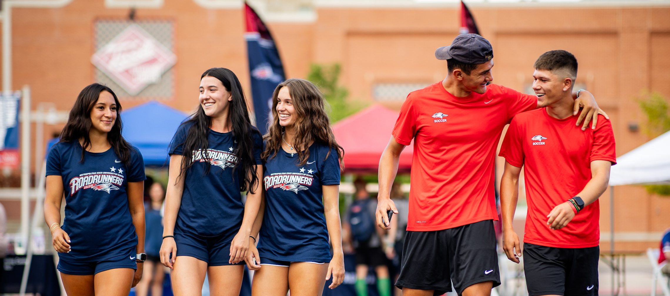 Five students walking together outside, looking at each other and smiling while wearing MSU Denver blue and red t shirts