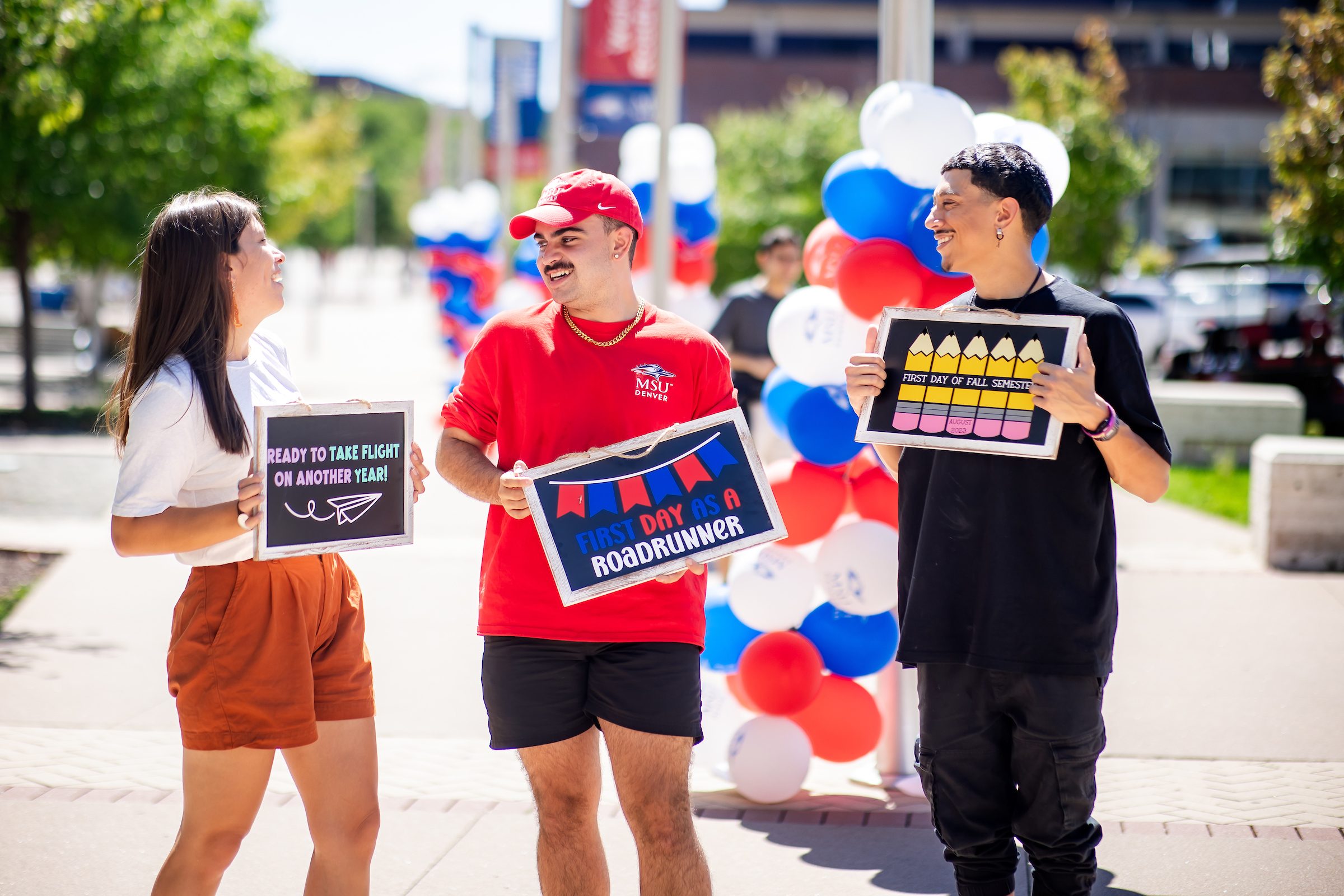 Three students together with signs welcoming students, smiling at each other during the first week of classes