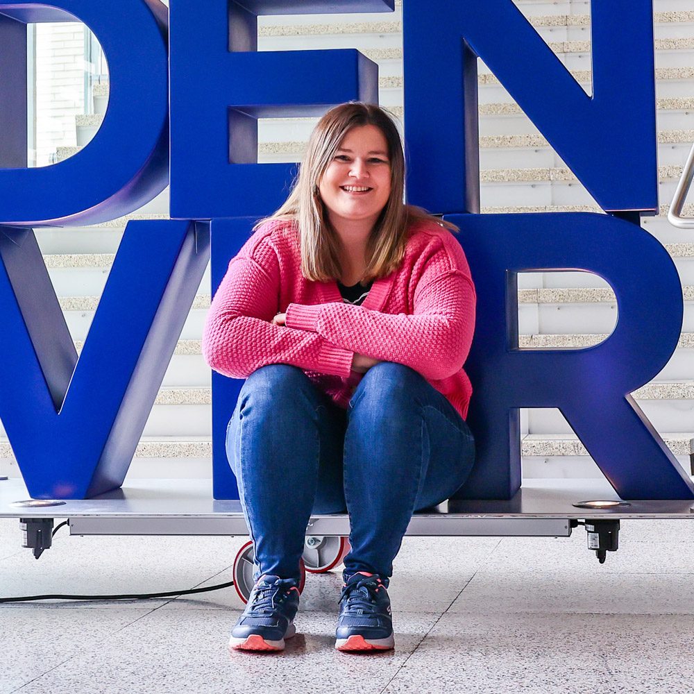 MSU Denver social work graduate student Christy Walker sits in front of the red and blue MSU Denver sign inside JSSB
