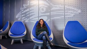 Aerospace engineering technology major Evelyn Montes Gonzalez sitting in front of the white Roadrunner birdhead in the JSSB student lounge in a blue chair
