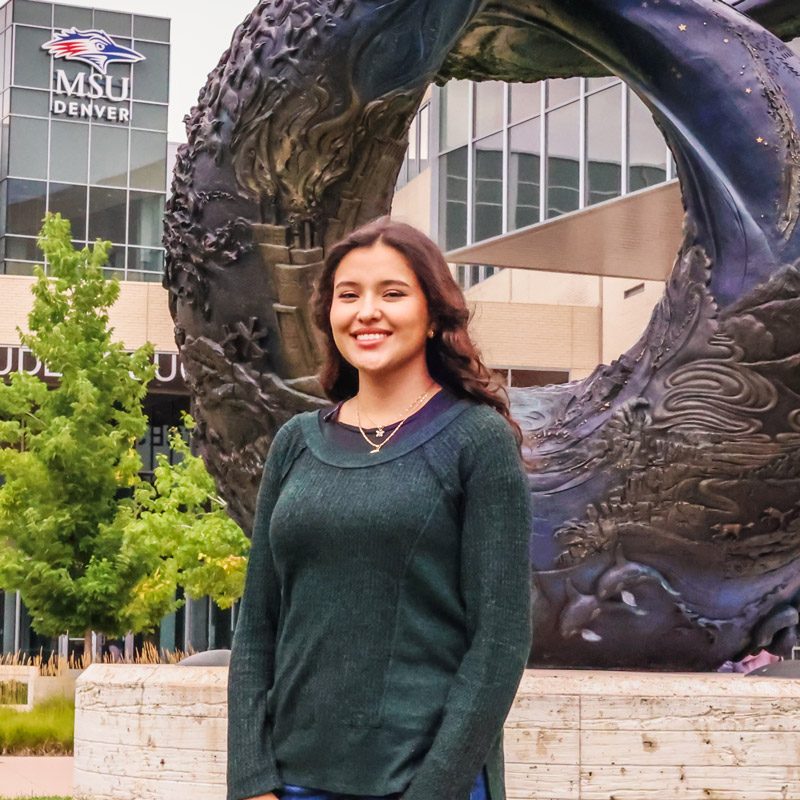 Aerospace engineering technology major Evelyn Montes Gonzalez standing in front of the One World, One Water statue in front of JSSB with the MSU Denver sign in the background