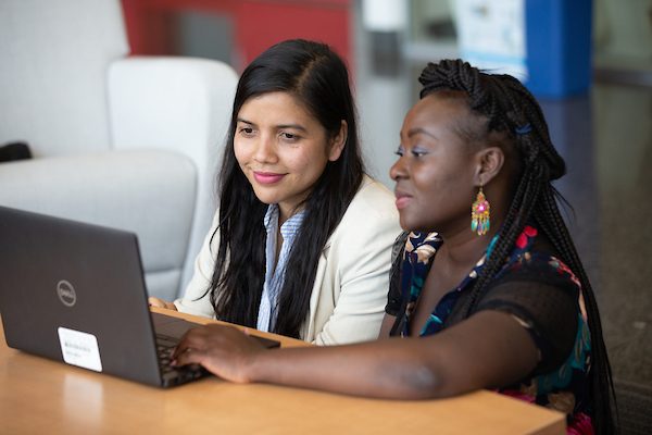 Two young woman sit side by side, looking at a laptop screen