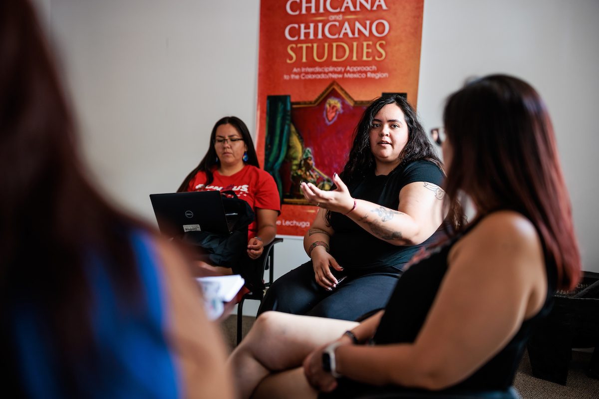 Three people sitting and talking with a Chicana and Chicano Studies book cover in the background