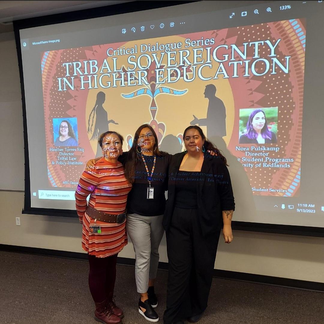 Three Native American staff members pose in front of an event flyer. Event Flyer text: Tribal Sovereignty in Higher Education