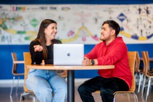 A male and female student talking and smiling at a table with a laptop
