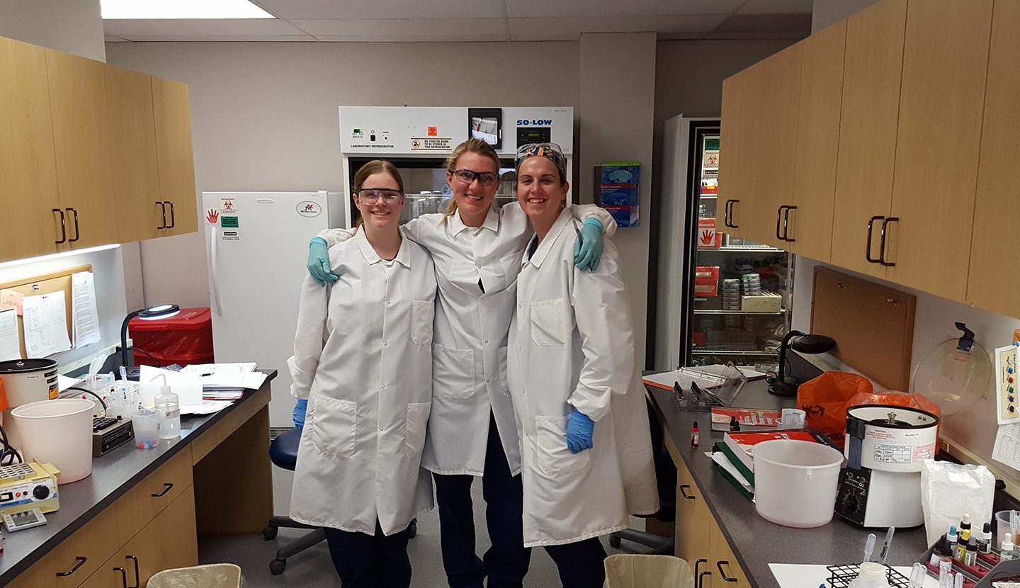 Three women wearing lab coats standing together in classroom lab