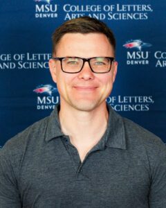 A smiling man wearing glasses and a gray collared shirt stands in front of a blue backdrop featuring the MSU Denver College of Letters, Arts and Sciences logo.