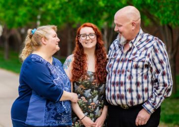 A female graduate standing with her parents outside on campus at First Generation Graduation