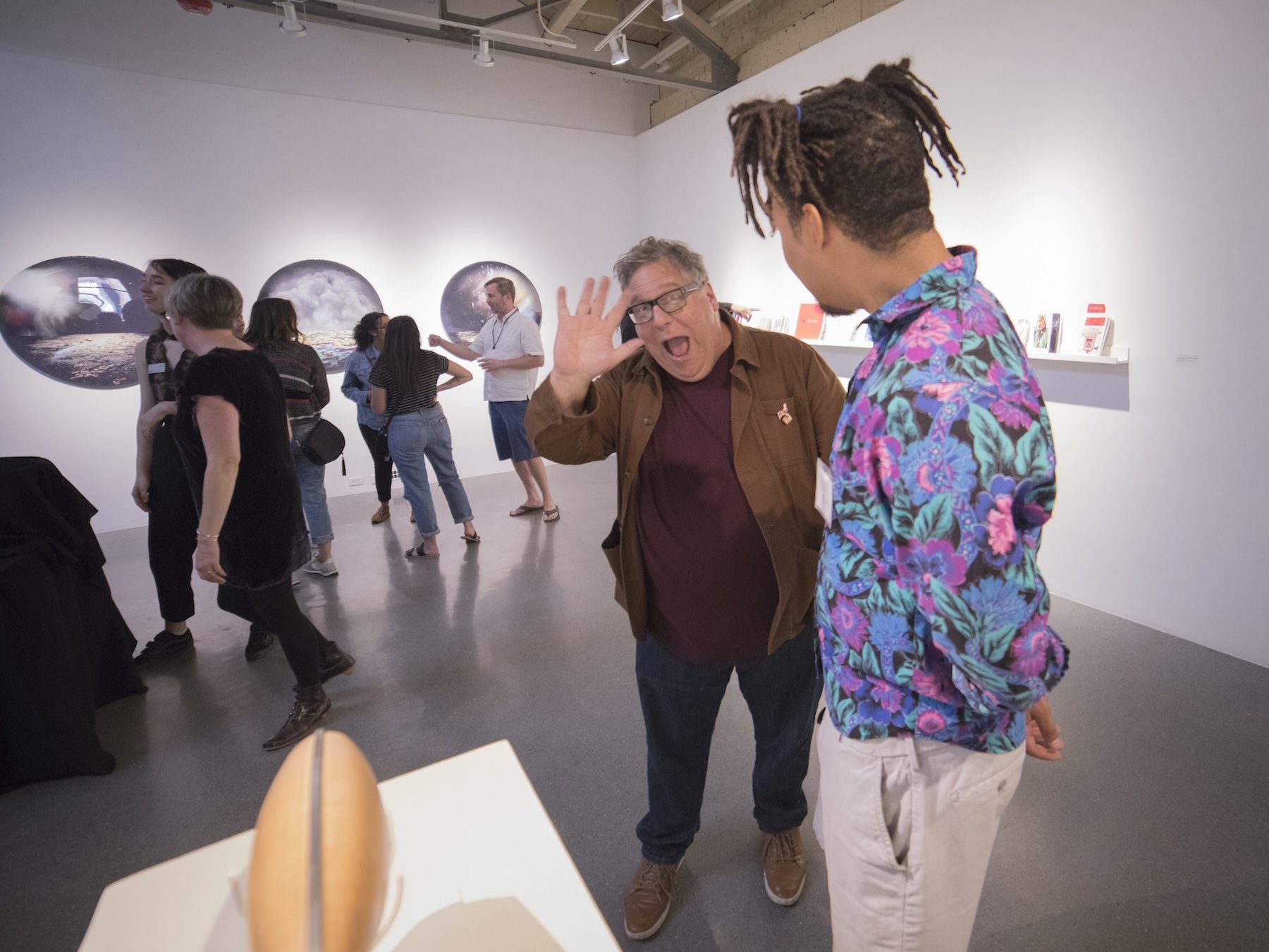 A guest and an artist interacting in the gallery space, with the two main subjects in the foreground. One is wearing a bright blue shirt with purple floral patterns, while the other gentleman is waving at the camera.
