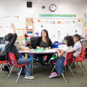 Photograph of a female elementary teacher sitting at a table with 4 young students, one of whom has their hand raised.