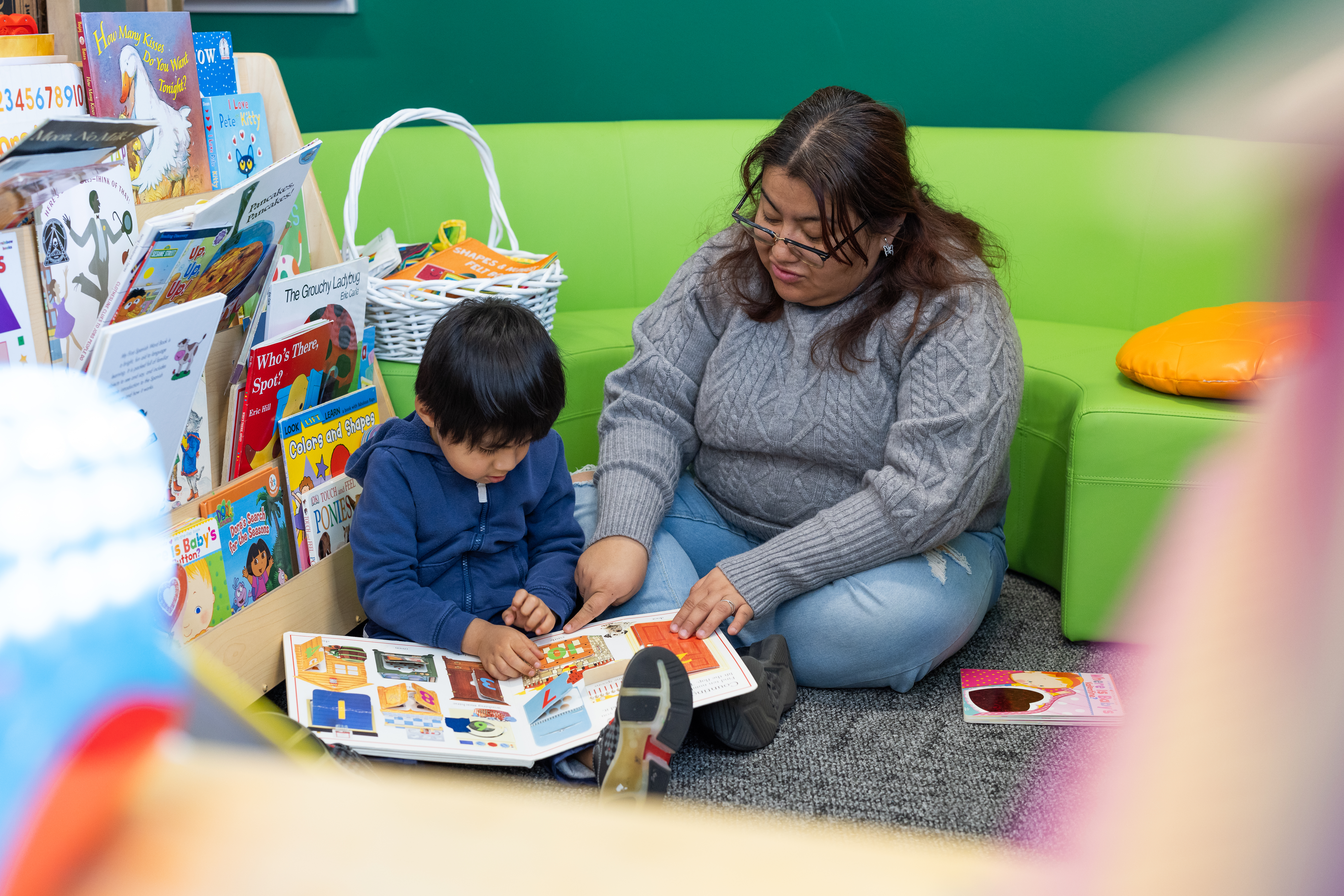 A woman sits on the floor with a young child reading a book next to a rack full of children's books.