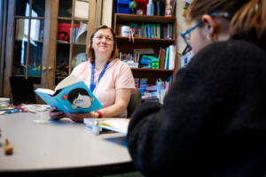 Photo of a teacher sitting at a table with a book, looking up and smiling at a student.