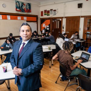 Photograph of a male teacher wearing a suit standing in a classroom with students sitting at tables working on assignments.