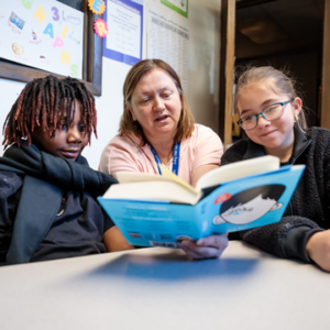 Photograph of teacher sitting at a table with two students reading a book.