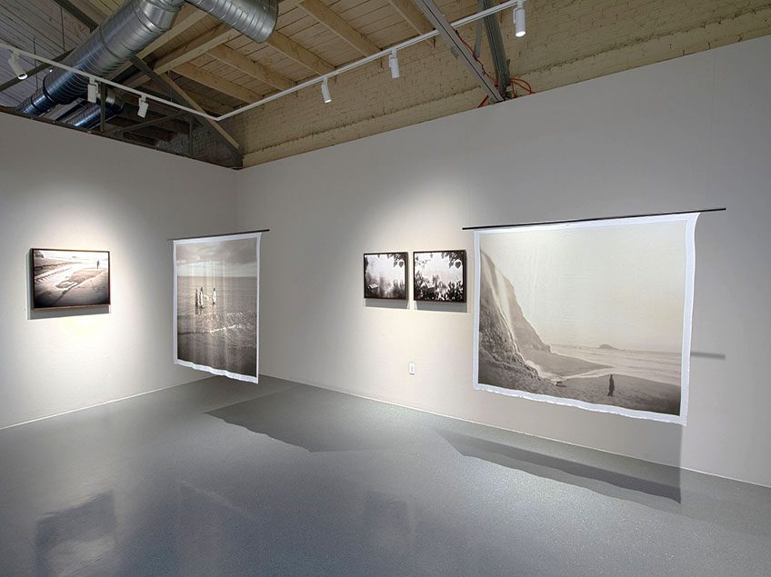 Photograph of the art gallery. On the walls there are small rectangular framed photographs of things like beaches, water, and a floating urn. There are silk photographs hanging.