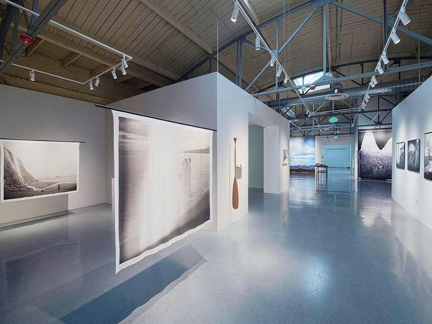 Photograph of the gallery space during Tomiko Jones: The Intimate Infinite. In the foreground is hanging silk rectangles with black and white photographs on them. In the background, you can see a glimpse of the rest of the gallery, where there are large photographic murals and framed art.