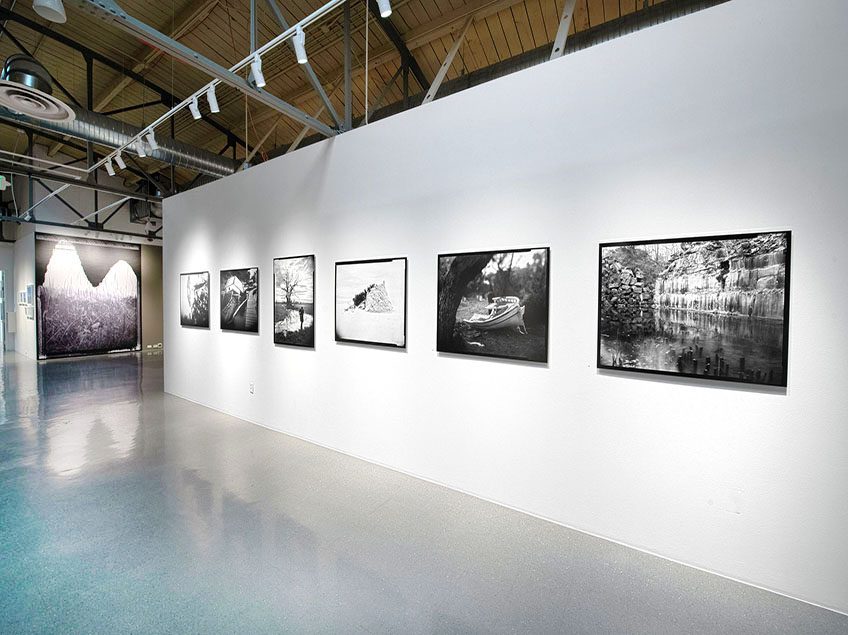 Six black and white photographs hang on a white wall. The subjects vary, from natural landscapes to boats. In the background, you can see the glimpse of a black and white floor-to-ceiling mural.
