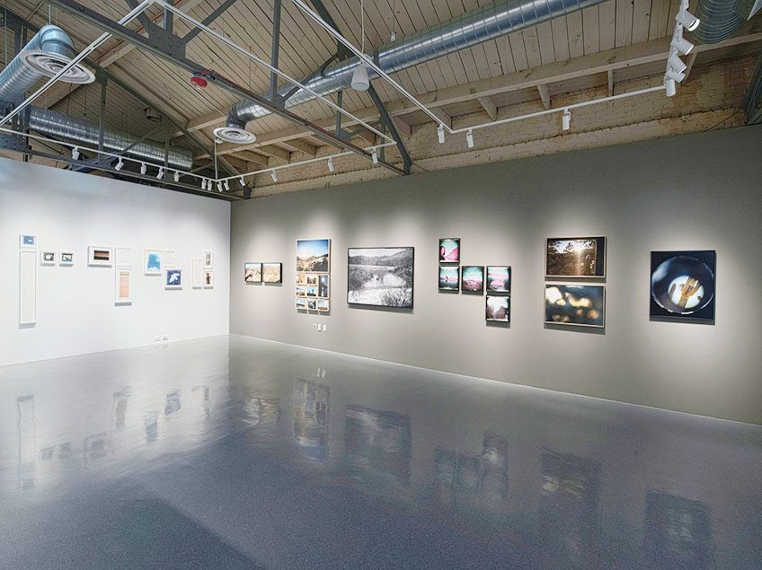 Art gallery interior with framed artworks on white and gray walls under exposed wooden ceilings.