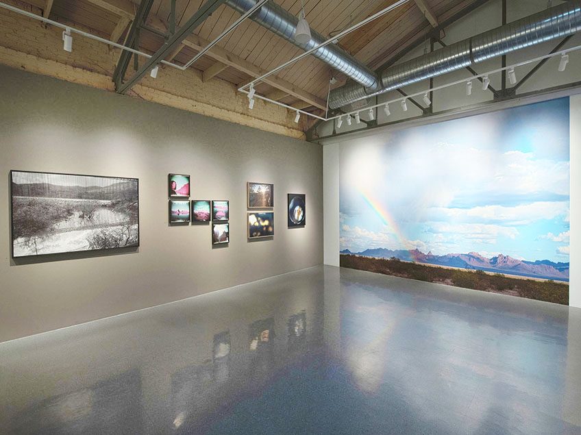 Framed multicolored and black and white photographs hang on a grey wall on the left of the photo. On the right, there is a photographic mural of a sky and rainbow in the desert.