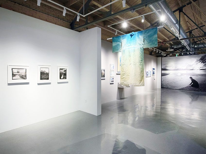 Photograph of gallery space at CVA. On the left wall there are framed photographic prints. Hanging in the middle of the gallery is a blue and yellow yukata. At the end of the hallway is a large black and white mural of a woman kneeling at the edge of a lake, where there are snowy mountains in the background.