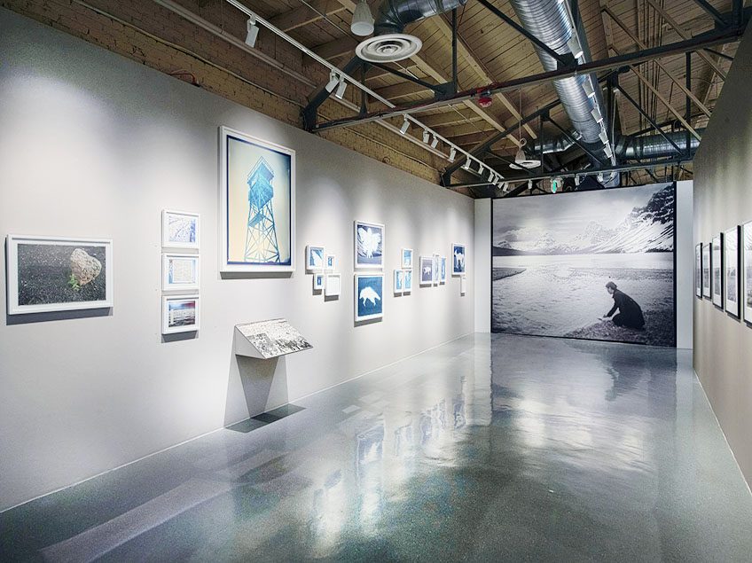 Photograph of gallery space at CVA. On the left wall there are many framed cyanotype prints. On the right wall there are black and white framed photos. At the end of the hallway is a large black and white mural of a woman kneeling at the edge of a lake, where there are snowy mountains in the background.