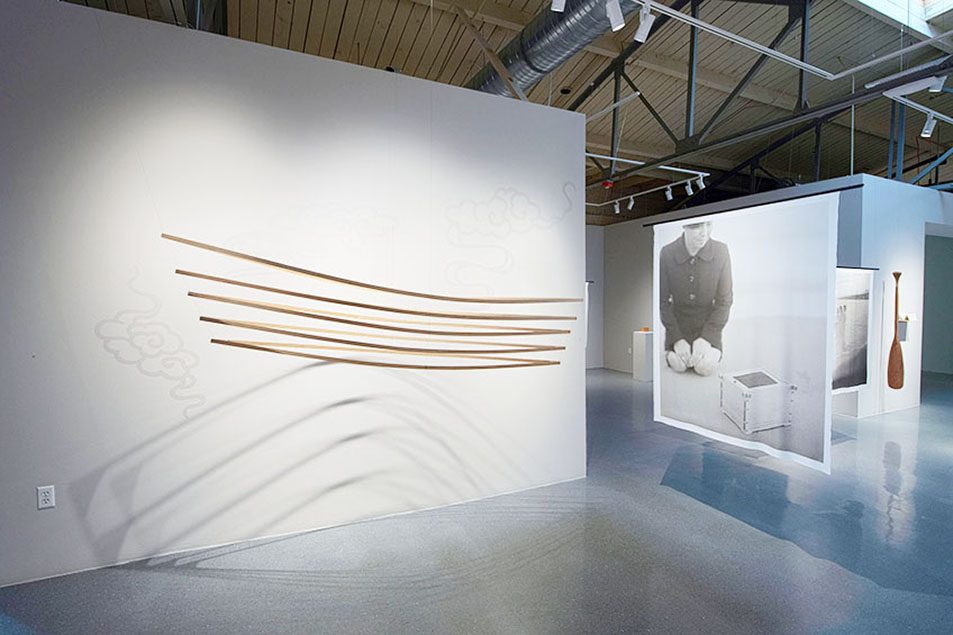 Hanging in front of a wall is the small frame of a wooden boat. On the right, there are hanging sheets of silk which have black and white photographs printed on them. The most prominent one has a woman kneeling next to a wooden box.