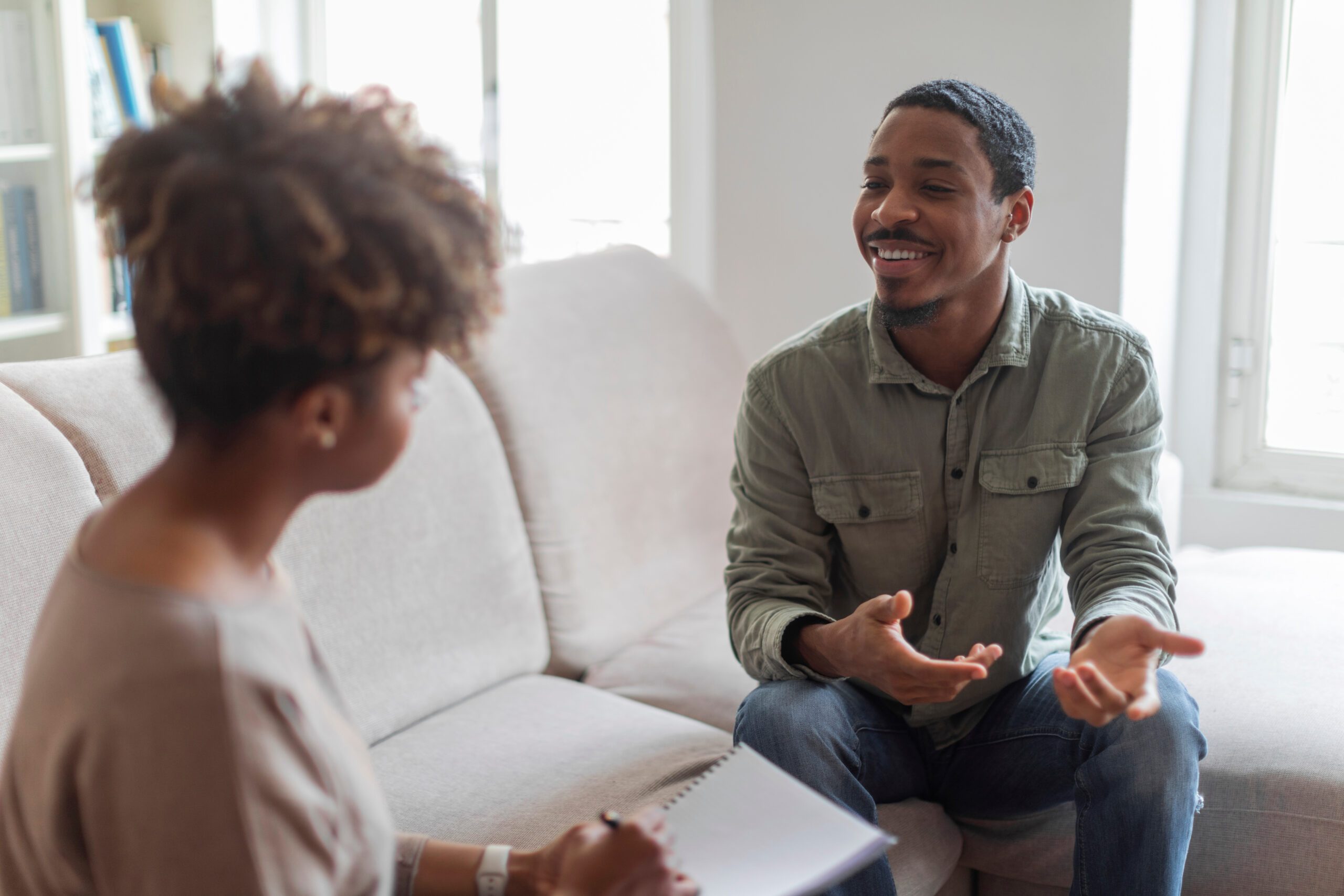 Cheerful positive handsome young african american guy in casual outfit sitting on couch at cozy counselor office, having conversation with therapist black woman, sharing good results of therapy
