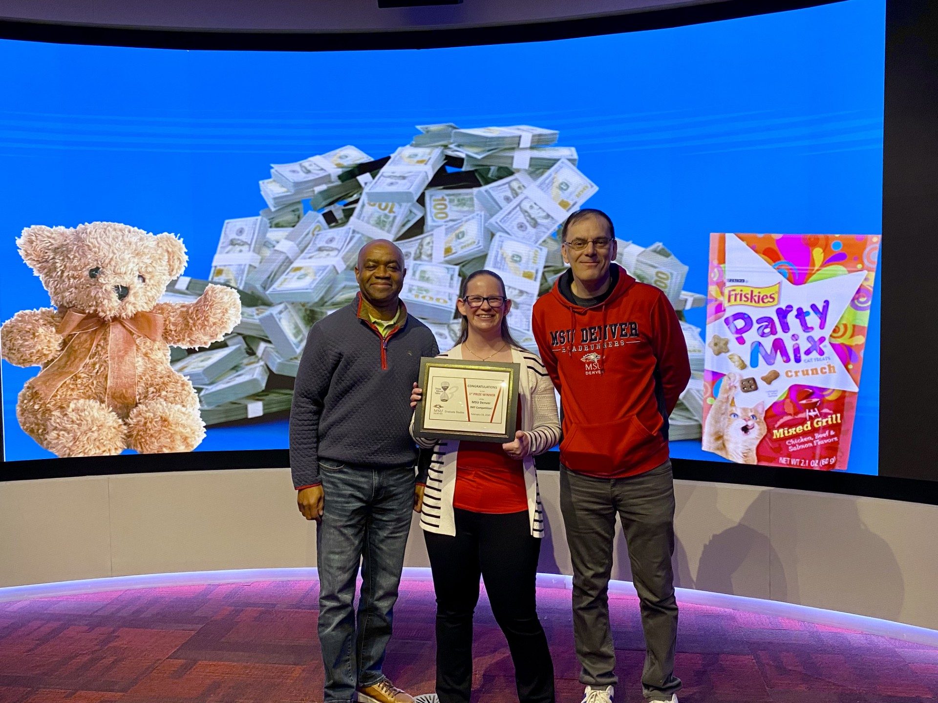 MPAcc Alum Laura Girdner places 1st at the MSU Denver 3MT final competition - stands with Acc Department Chair, Dr. Clifton, and Faculty Advisor, Dr. Andrew Holt, with certificate in front of her slide
