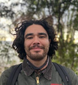 Head shot of a man with long hair and short beard with trees in the background