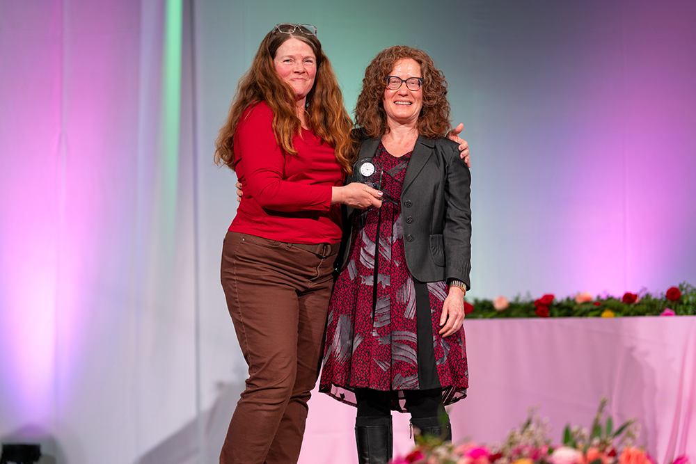Lisa Suter, professor of English, presents Elizabeth Kleinfeld, professor of English, with the inaugural Roadrunners Who Soar Scholarly or Creative Activities award. Both stand smiling on stage, with Elizabeth holding the award.