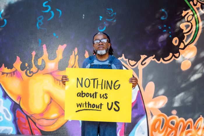 A Deaf Black man wearing glasses looks neutrally at the camera while holding a sign declaring 
