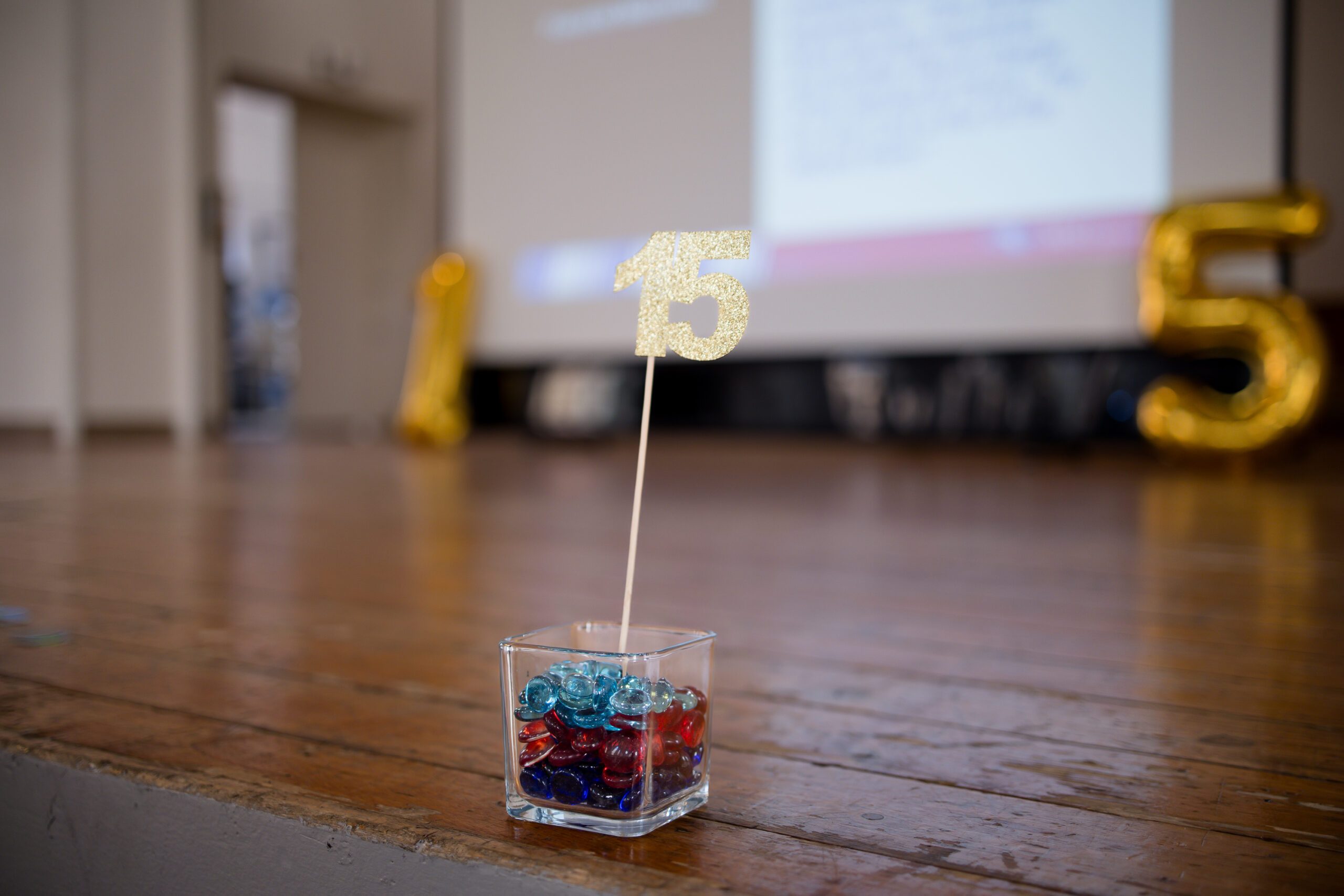 close up of some stage decorations at MPAcc's 15 year anniversary event - background of slide screen with a gold 1 balloon and gold 5 balloon on either side with a small square vase of red and blue marbles and a gold 15 standing right in front and focus