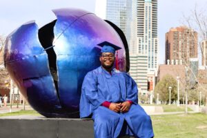 2025 MSU Denver graduate Chrisnel Akele sitting in front of a blue, round statue on campus