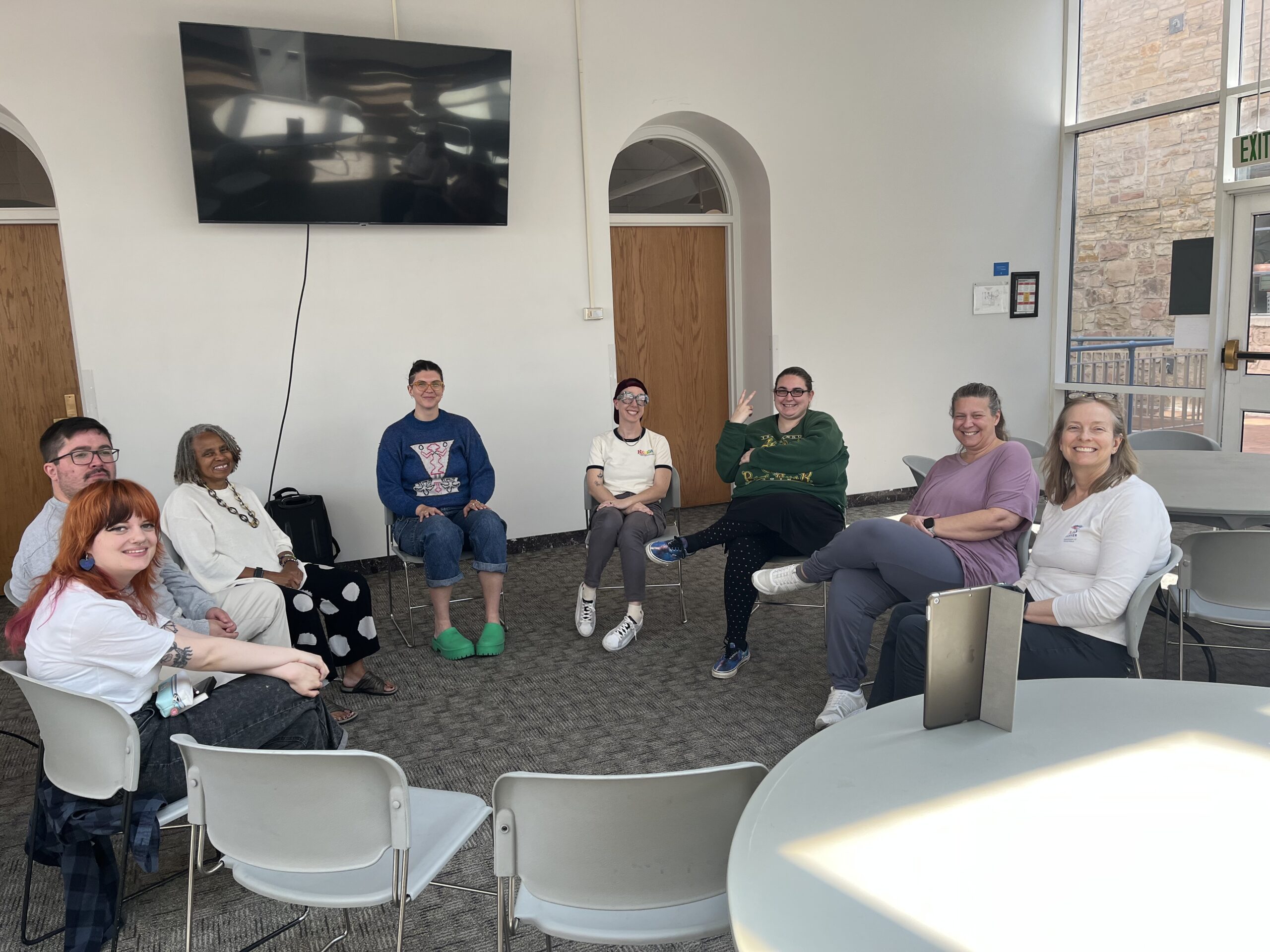A group of nine people sit in a circle of chairs in a bright room, smiling at the camera.
