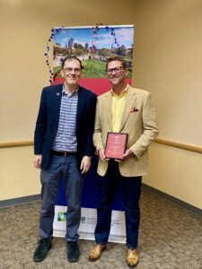 faculty Dr. Andrew Holt standing next to MPAcc recipient, Rory Yonts, holding red Outstanding Student Award plaque in front of MSU Denver sign