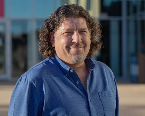 A man with shoulder length, wavy brown hair and wearing a blue button down shirt smiles for the camera.
