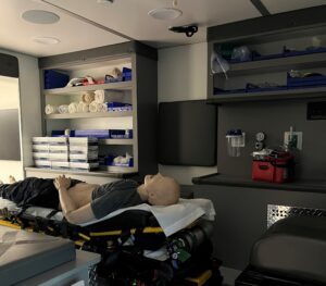 Photo is of the EMS Ambulance simulation with a simulation mannequin on a gurney surrounded by medical equipment.