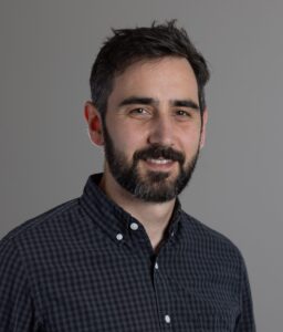 Professional headshot of a bearded man in a black button up with white stripes in front of a gray background