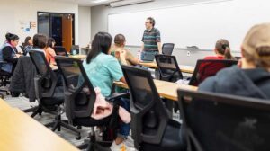 Professor lecturing at the front of a classroom in front of a whiteboard
