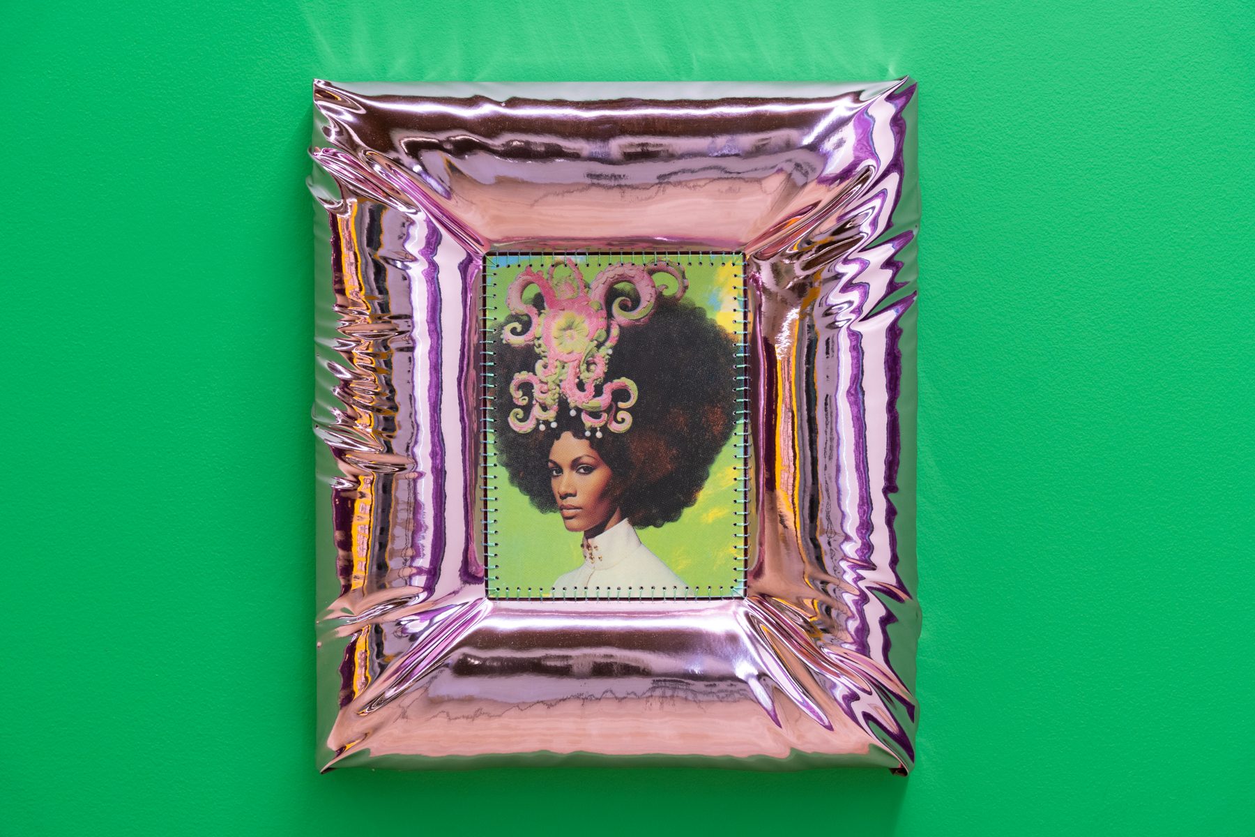 The image depicts a square artwork placed against a vivid green wall. The art piece is encased in a glossy, reflective frame that has a metallic, rose-gold hue with creased and crinkled edges, creating an illusion of movement and depth. The center of the frame holds an image of a person with an elaborate hairstyle featuring large curls and decorative, intricate designs in shades of pink and white against a bright green background. The contrast between the vivid backdrop and the metallic frame emphasizes the central portrait.