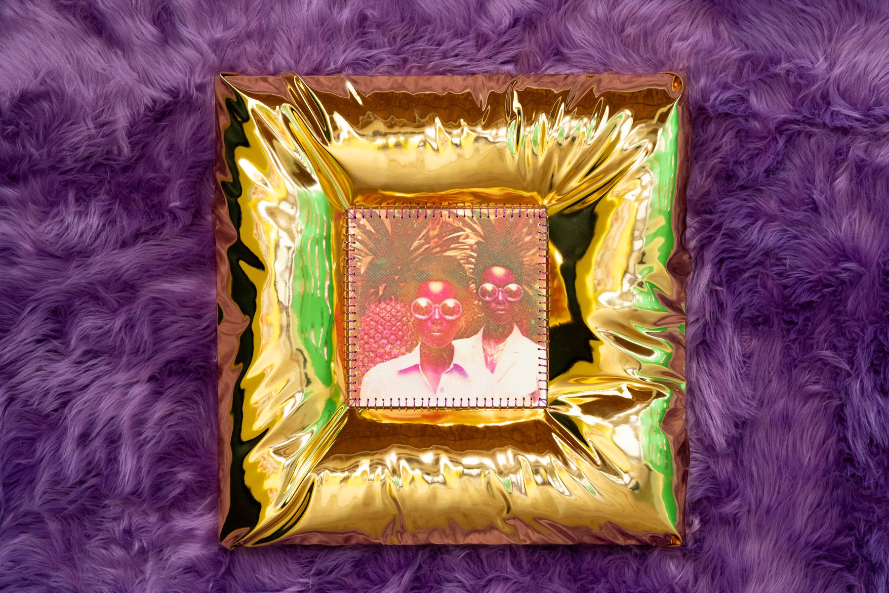 A photograph placed at the center of a shiny, gold metallic frame with an inflated appearance. The frame is square-shaped and reflects light, creating an iridescent effect. The photo within the frame depicts two individuals wearing large sunglasses and standing close together. They are surrounded by vibrant, tropical imagery, including pineapples. This framed photo is placed on a plush, textured purple fabric background that covers the entire visible area around the frame.