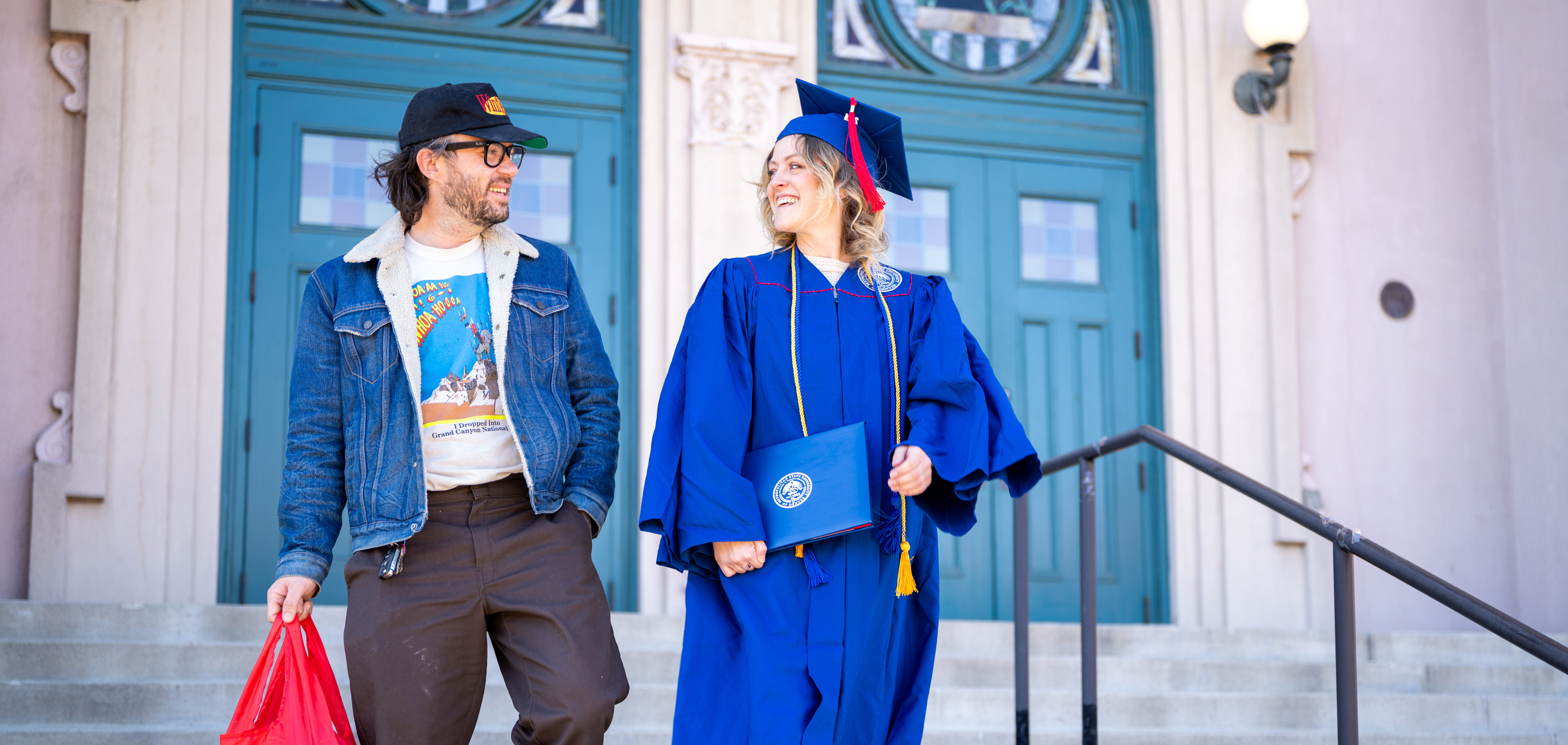 Graduate in cap and gown smiles at man in a hat holding an MSU Denver tote bag.