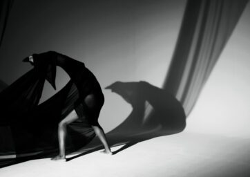 The black and white image depicts a figure draped in flowing black fabric, creating a dynamic and sculptural form against a stark, minimalistic backdrop.