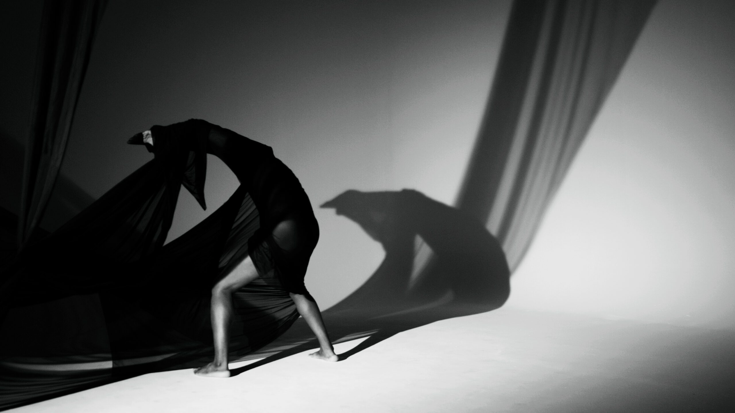 The black and white image depicts a figure draped in flowing black fabric, creating a dynamic and sculptural form against a stark, minimalistic backdrop.