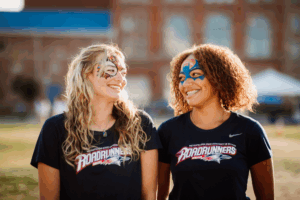 2 MSU Denver students looking at each other. They are wearing MSU Denver T-shirts and have face painting on their faces.