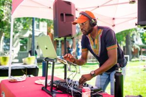 A DJ wearing headphones, sunglasses, a traditional African-style shirt, and an orange cap is performing at an outdoor event under a pink canopy. He is using a laptop and mixer on a red table with drinks, plates, and speakers around him. The setting is a sunny day on a green lawn with blurred people and trees in the background.