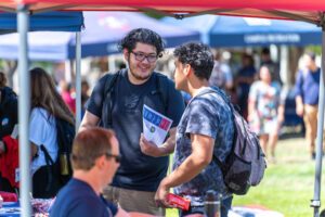 Two students talk and smile under a red tent during a campus fair, holding informational materials and wearing backpacks. Tables with flyers and staff are in the foreground, with other booths and attendees visible in the background on a sunny day.