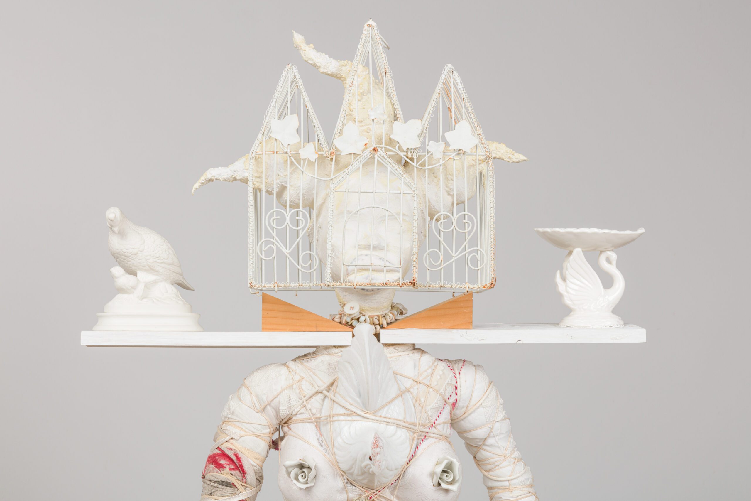 Sculpture of a wrapped torso with a cage-like structure as a head, flanked by porcelain objects.